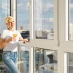 Positive middle-aged woman on balcony of high-rise apartment with glass of juice under bright sun