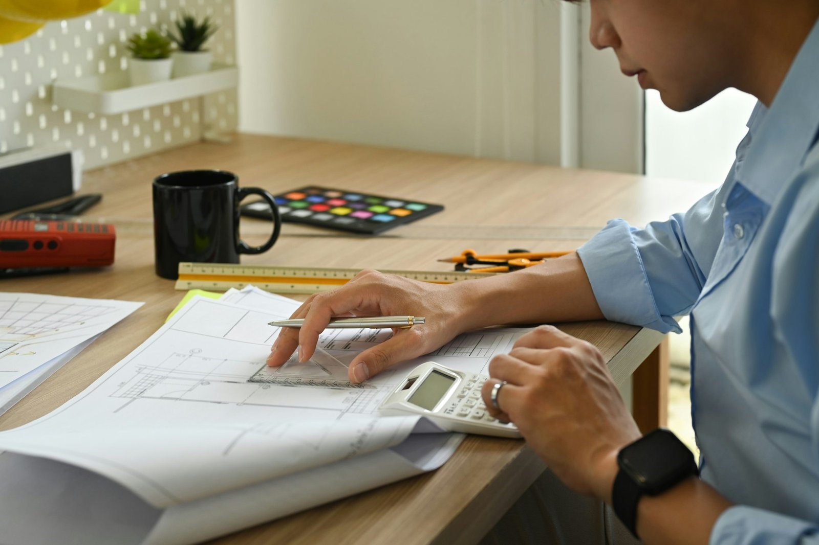 Cropped shot of engineer using calculator, examining construction plans at his workstation.