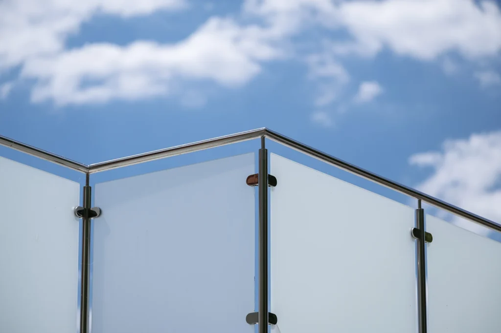 Glasräcke med rostfria stolpar och frostat glas monterat utomhus mot en blå himmel med moln.